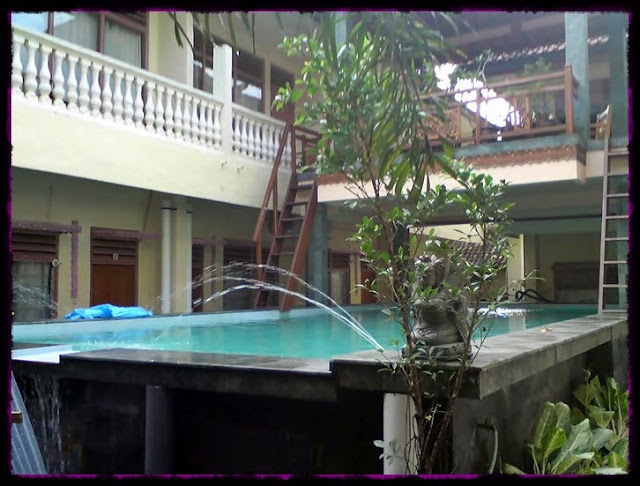Pemandangan halaman dalaman sebuah bangunan dua tingkat dengan balkoni, kolam kecil berpancutan, dan kehijauan di sekitarnya dalam warna-warna lembut.