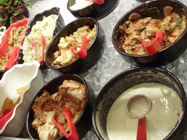 Pemandangan dari atas beberapa mangkuk gelap berisi pelbagai hidangan sampingan dan salad di atas lapisan ais dengan penyepit merah.